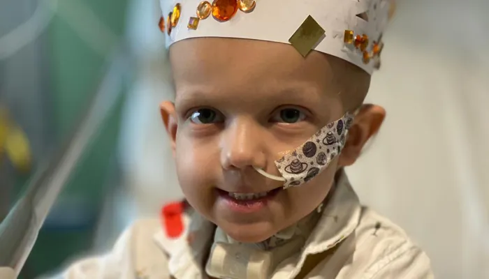 A photo of a smiling young boy sitting on a hospital bed with a tube in his nose, wearing a white paper crown decorated with shiny bits of paper.