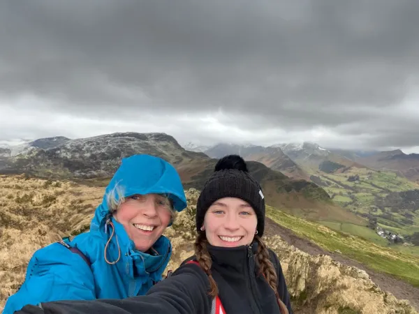 Two smiling young women wearing walking clothes are standing in front of a green landscape, with dark clouds above them