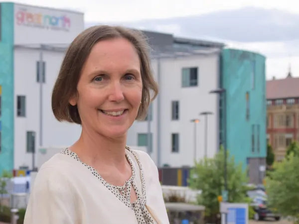 A woman with short brown hair is wearing a cream cardigan and standing in front of a children's hospital