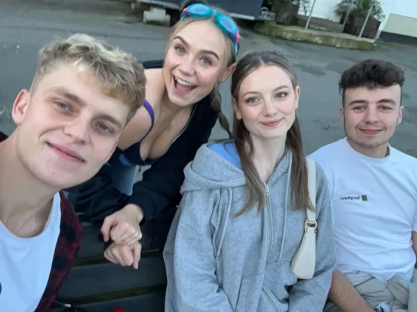 A group of four young friends smile for a photo. The group includes two girls and two boys: one girl is blonde with blue swimming goggles on her head, and the other is a brunette wearing a grey hoodie. One boy is blonde, wearing a white t-shirt and a dark red jacket, while the other is brunette, wearing a white t-shirt.