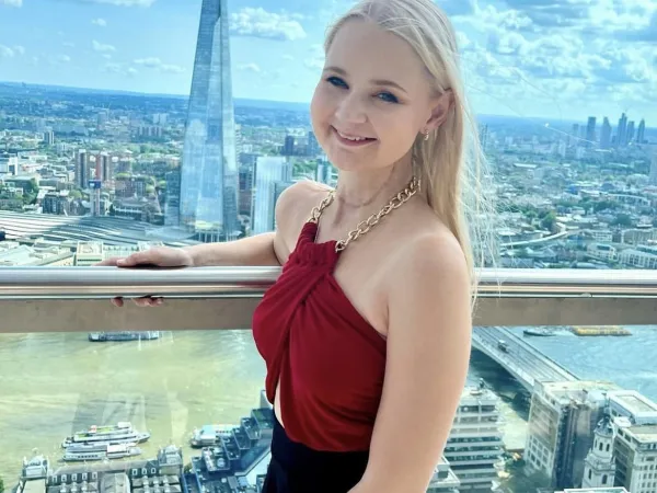 A young blonde woman stands by a window in a tall building, overlooking the city of London. She wears a stylish red top and a black skirt.