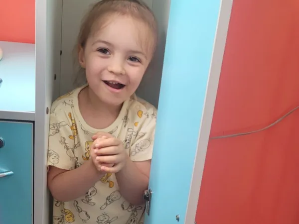 A little girl wearing a yellow t-shirt is playing, hiding inside a blue and red cupboard with the door open, smiling and holding her hands together.