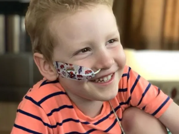 A young boy with a nasogastric tube is smiling while wearing a striped orange t-shirt.