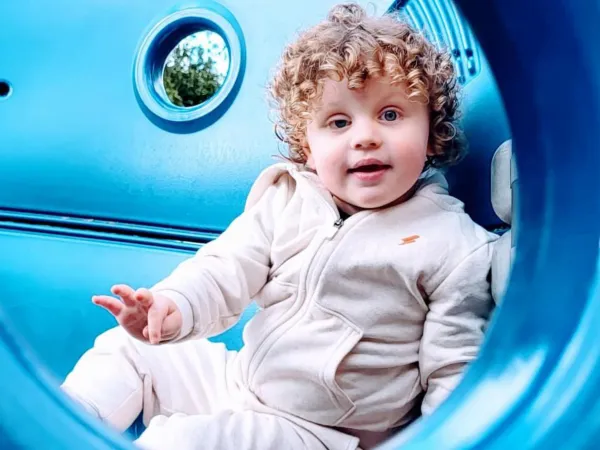 A smiling young boy with curly brown hair is sitting inside a blue climbing frame, wearing a grey tracksuit that matches.  