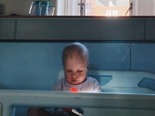 A toddler wearing a light blue t-shirt sits in a hospital bed, with his head down. 