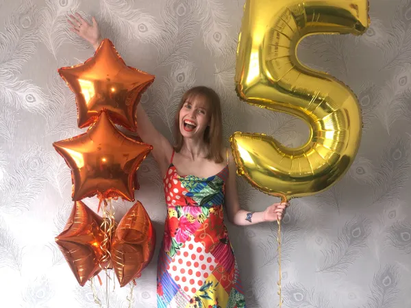 A young girl wearing a bright coloured dress is holding a number '5' gold balloon an d four orange heart balloons.