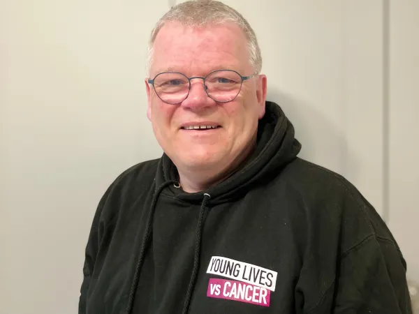 A man is standing against a white background, wearing glasses and a black hoodie with the logo of Young Lives vs Cancer.