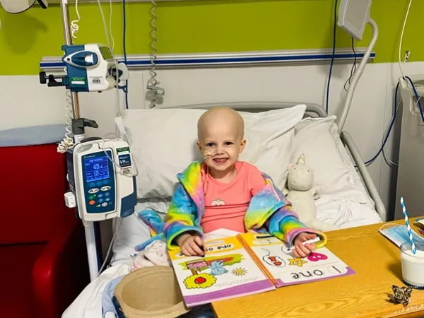 A young girl with a bald head sits in a hospital bed, smiling. She wears a colourful rainbow-patterned dressing gown, with a bright colouring book open in front of her.