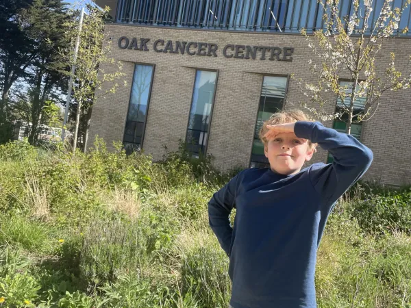 A young boy with curly blonde hair is happily standing outside the Oak Cancer Centre.