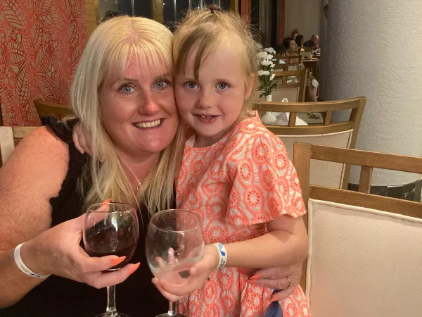 A mother and young daughter sit closely at a dinner table, arms around each other, smiling and holding drinks.