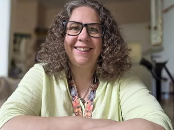 A woman with curly brown hair, glasses, and a yellow top sits with her arms crossed, smiling at the camera.
