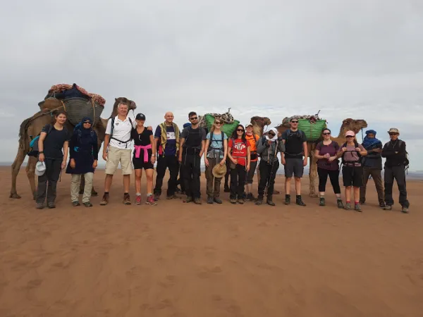 A group of people with rucksacks and three camels behind them standing in the sahara desert