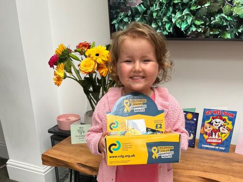 Arabella, a young girl who was diagnosed with acute lymphoblastic leukaemia, smiles at the camera whilst holding a CCLG Childhood Cancer Awareness Month appeal box.