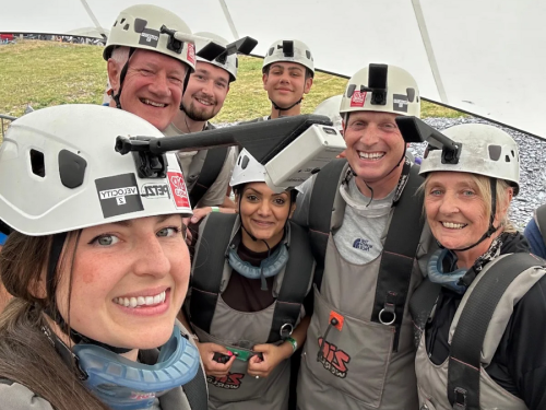 A group of 6 smiling people, all wearing helmets and safety gear for doing a zip line challenge, are taking a group selfie.