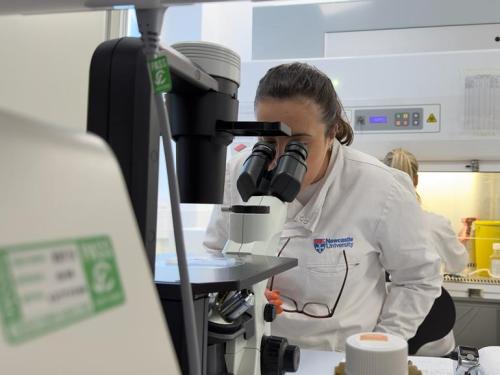 A woman in a research lab, in a white lab coat, looking into a large microscope.