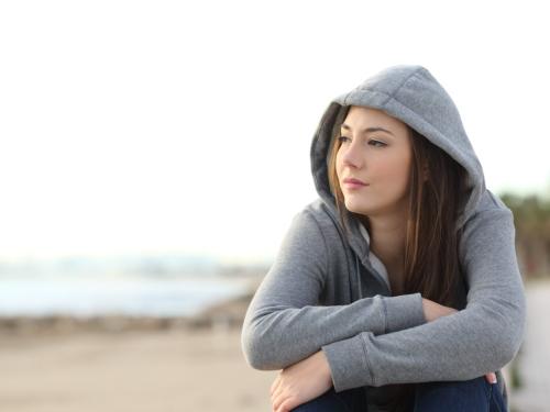 A young woman in a grey hoodie, sitting on a beach and looking thoughtful.