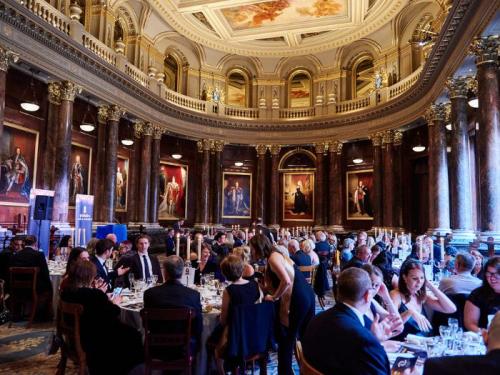 Tables of people in black tie attire in a large, ornate dining room as part of a gala event
