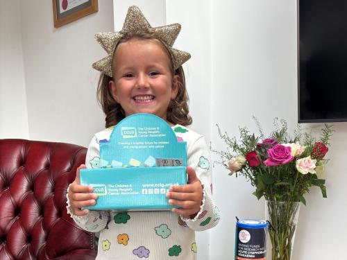 A young girl in spotty pyjamas wearing a star headband, smiling and holding a CCLG badge box in front of her