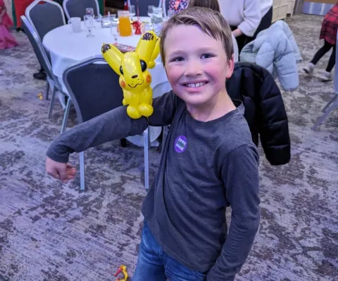 A young boy in a long-sleeve navy top is smiling at the camera with his arm out balancing a yellow balloon character.