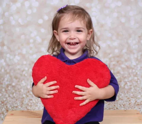 A young girl with brown hair smiles at the camera, wearing a purple top and holding a large red fluffy heart.