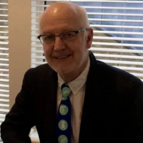 A man with grey hair and glasses is dressed in a white shirt and a black blazer. He is wearing a blue tie with lighter blue circles.