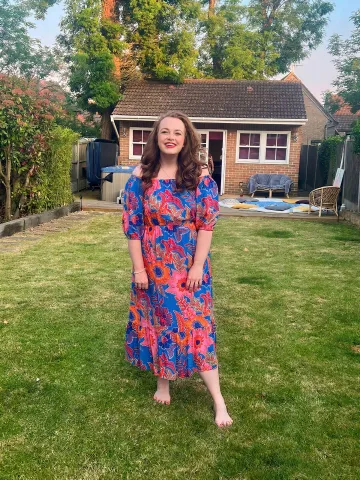 A young woman in her 20s with long brown hair is standing in a garden, smiling at the camera. She is wearing a red and blue floral dress.