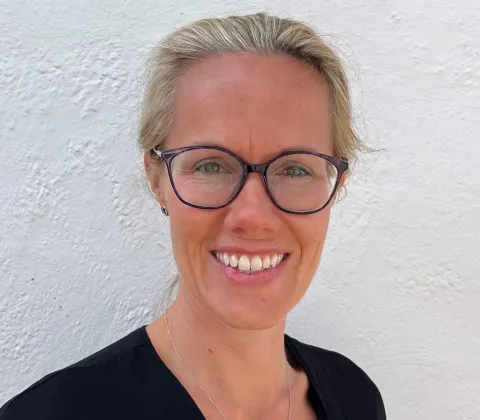 A headshot of a smiling blonde woman wearing glasses and a black shirt, taken in front of a white wall.