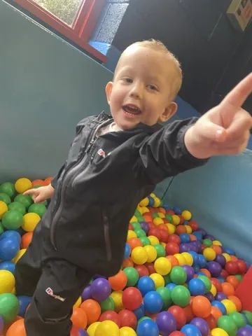 A young boy with short blonde hair is playing in a colourful ball pit, smiling and pointing past the camera.