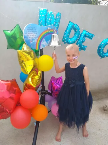  A young girl with a bald head and a nasogastric tube is ringing the End of Treatment Bell. She is wearing a navy party dress, surrounded by colourful balloons and decorations.