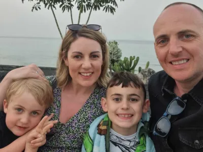  A family consisting of a mum, dad and their two children smiles at the camera, with the ocean and trees in the background.