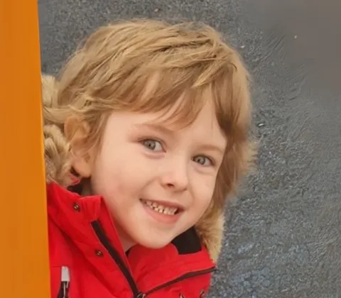 A young boy smiles at the camera while peeking around an orange climbing frame.