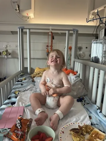 A young girl wearing a nappy is sitting upright in a hospital cot, surrounded by various medical equipment.