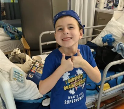 A young boy wearing a blue hat and t-shirt is leaning against a hospital bed, giving a thumbs up to the camera.