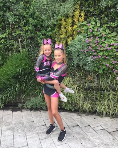 Two girls stand together in pink and black cheerleading uniforms. The older girl is holding the younger girl up.