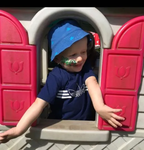 A young boy with a nasogastric tube is wearing a blue hat while leaning out of a plastic playhouse window.