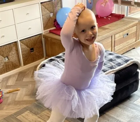 A young girl with a bald head is wearing a purple tutu, dancing like a ballerina.