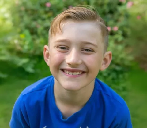 A smiling young boy with short blonde hair is standing in a sunlit garden, wearing a bright blue t-shirt.