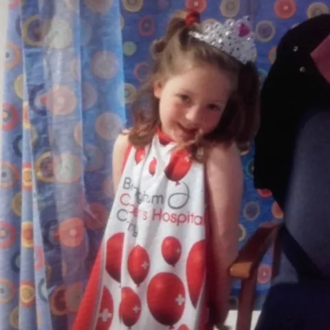 A young girl with long brown hair in pigtails is smiling in front of a hospital curtain, wearing a Birmingham Children's Hospital gown. 