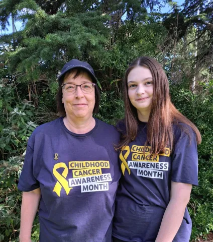  A mother and daughter stand close together, wearing matching navy blue t-shirts with a Childhood Cancer Awareness Month logo on.