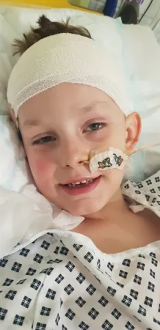 A young boy lies in a hospital bed with a head bandage and a nasogastric tube on his face..