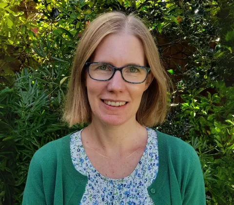 A woman with shoulder-length hair and glasses is standing in front of a green hedge. She is wearing a floral T-shirt and a green cardigan. 