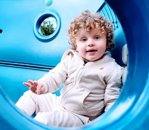 A smiling young boy with curly brown hair is sitting inside a blue climbing frame, wearing a grey tracksuit that matches.  