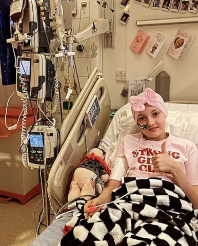 A young girl wearing a pink headband and a t-shirt that says "Strong Girls Club" is lying in a hospital bed surrounded by medical equipment.