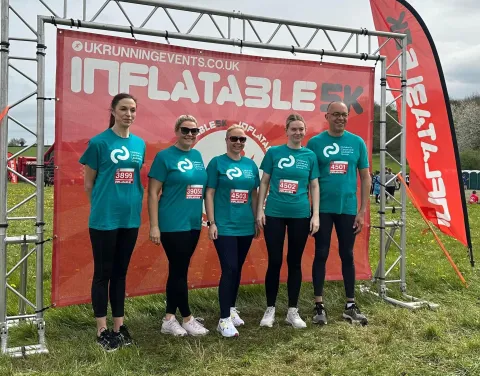 A group of five individuals, consisting of four women and one man, stands together in front of a red inflatable background marking the start of a 5K race. They are all wearing matching teal t-shirts with the CCLG logo.