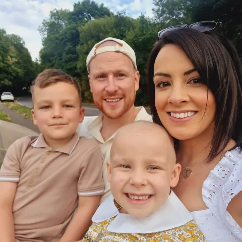 Woman smiling and facing camera with her husband and two children. One boy and one girl. The little girls has no hair and has cancer 