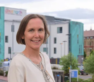 A woman with short brown hair is wearing a cream cardigan and standing in front of a children's hospital