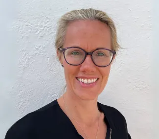 A headshot of a smiling blonde woman wearing glasses and a black shirt, taken in front of a white wall.