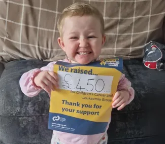 A young girl with short blonde hair is smiling as she holds up a yellow and navy poster featuring the CCLG logo. The poster displays the message: "We raised... £450 for Children's Cancer and Leukaemia Group. Thank you for your support." She is dressed in a pink long sleeve top.