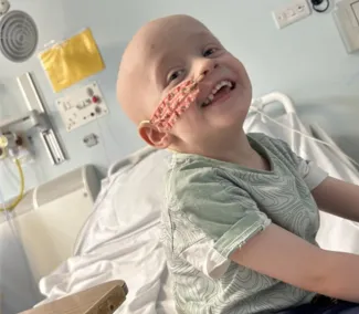 A young boy is smiling while sitting in a hospital bed, wearing a light green t-shirt. He has no hair and has a nasogastric tube fitted.