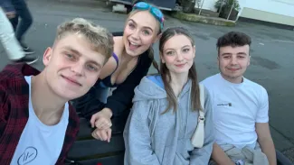 A group of four young friends smile for a photo. The group includes two girls and two boys: one girl is blonde with blue swimming goggles on her head, and the other is a brunette wearing a grey hoodie. One boy is blonde, wearing a white t-shirt and a dark red jacket, while the other is brunette, wearing a white t-shirt.